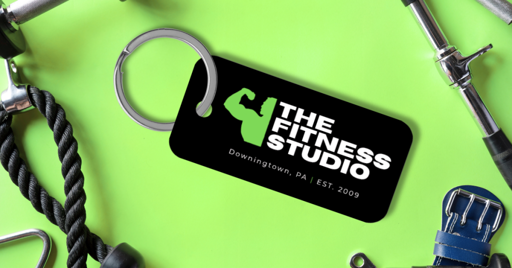 Black gym key tag with “The Fitness Studio” logo on a neon-green background, surrounded by fitness equipment.
