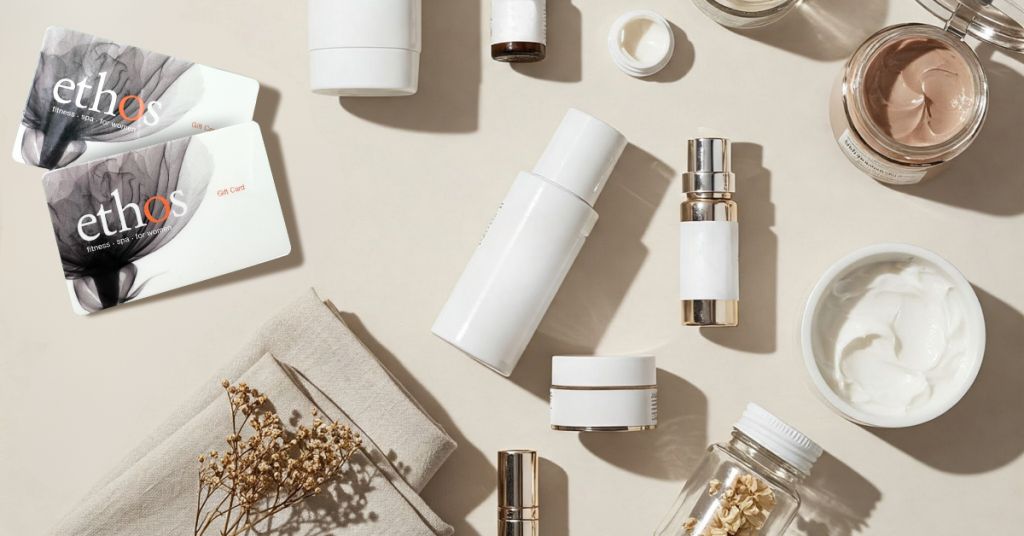 Flat lay of minimalist skincare and beauty products in white containers arranged on a beige background, including jars, pump bottles, droppers, and creams, alongside dried flowers, folded fabric, and two spa gift cards.