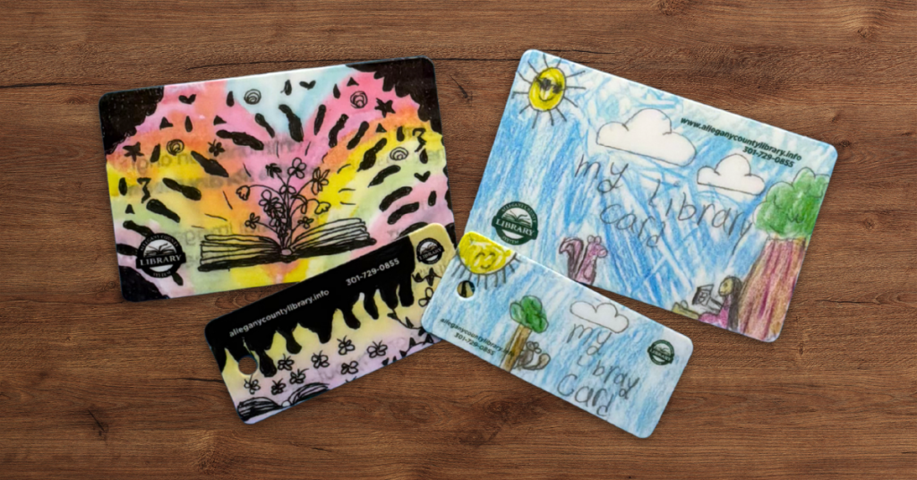 Colorful library card designs from Allegany County Library System, featuring hand-drawn illustrations of books, nature scenes, and abstract patterns displayed as printed cards and key tags on a wooden surface.