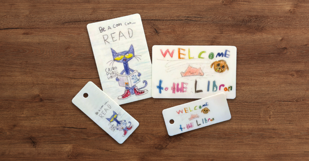 Hand-drawn library card designs from Cairo Public Library featuring a playful reading cat and colorful “Welcome to the Library” text, displayed as printed cards and key tags on a wooden surface.