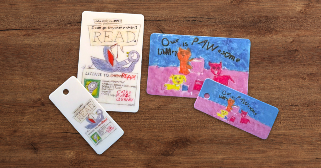 Hand-drawn library card designs from Cairo Public Library, featuring colorful illustrations of reading scenes and animals, displayed as printed cards and key tags on a wooden surface.