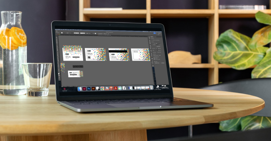 Laptop displaying gift card designs in Adobe software on a wooden desk workspace.