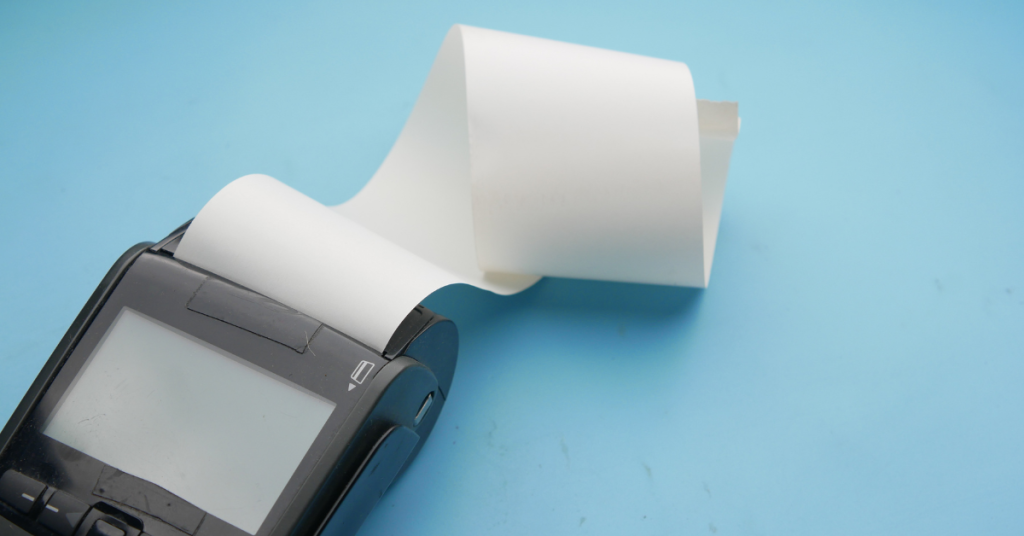 The image shows a small point-of-sale device with a roll of blank receipt paper feeding out of it. The device is black with a display screen, and it’s placed on a light blue background.